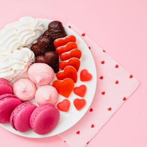 A top-view of a dessert platter with macarons, meringues, and heart-shaped candies on a pink background.