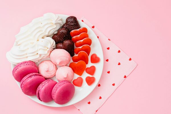 A top-view of a dessert platter with macarons, meringues, and heart-shaped candies on a pink background.